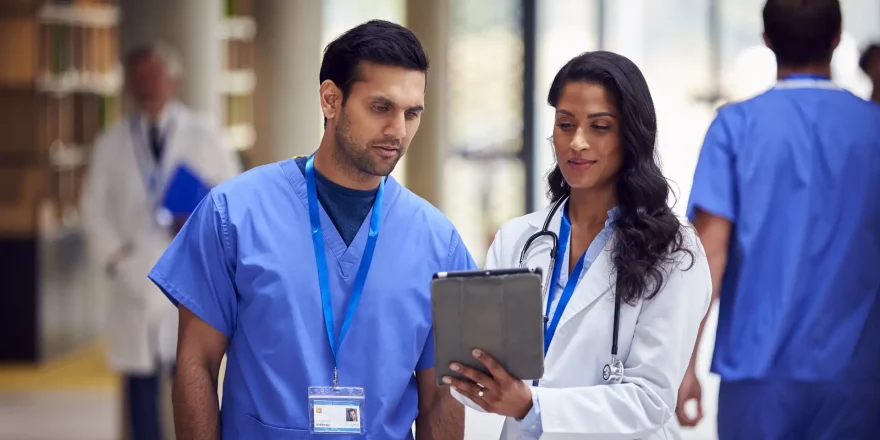 Two Medical Staff In White Coats And Scrubs With Digital Tablet Having Informal Meeting In Hospital